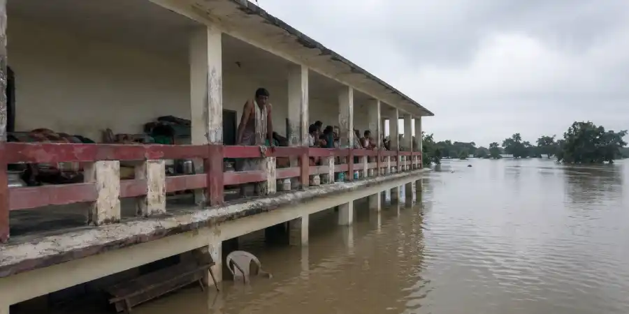 बाढ़ में डूबी स्कूल की निचली मंज़िल, ऊपर की बालकनी पर बैठे लोग और पानी में तैरता सामान