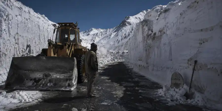 बर्फ़ काटकर बनाई गई ऊँची दीवारों के बीच पहाड़ी सड़क, किनारे पर डोज़र और किलोमीटर का पत्थर